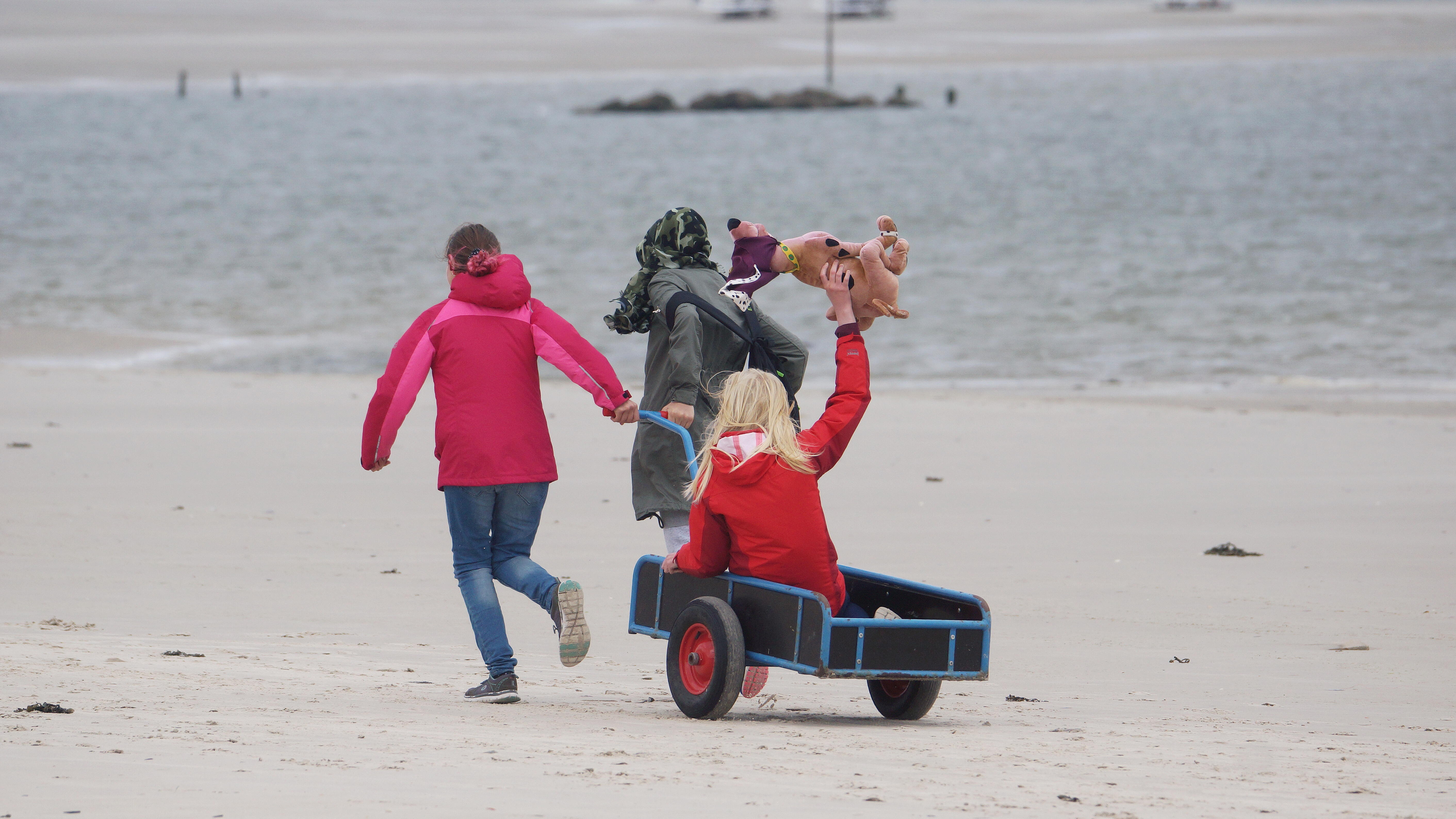 Foto: Strand und Nordsee. Zwei Kinder ziehen einen Wagen, in dem ein Kind sitzt. Rückenansicht.